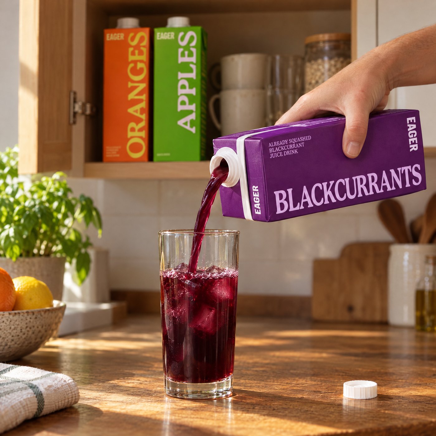 A hand pours dark purple juice from the Blackcurrants carton into a glass of ice on a wooden counter, with Orange and Apple cartons on an open cupboard shelf behind.